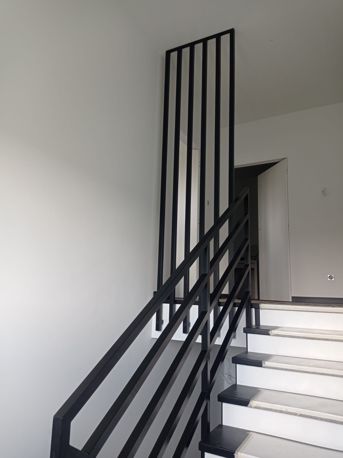 Nowoczesne schody z czarną metalową balustradą o minimalistycznym designie, z pionowymi szczeblami i białymi stopniami, prowadzące do otwartych drzwi w jasnym wnętrzu.