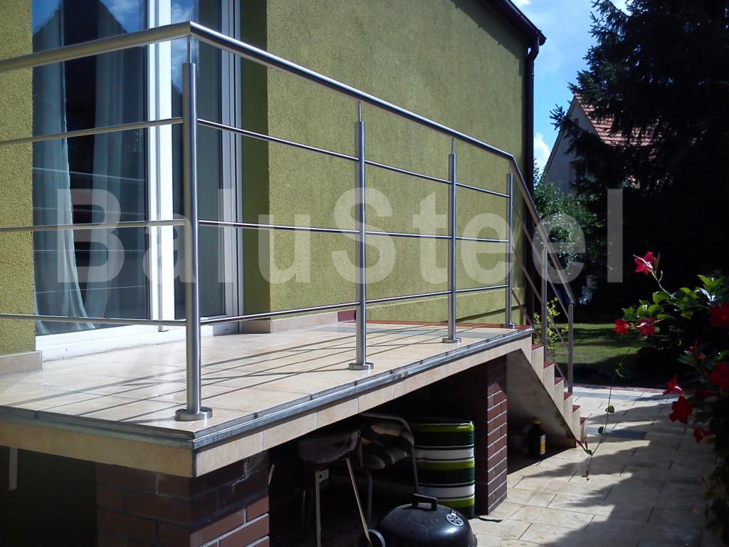 Zewnętrzna platforma z wyjściem balkonowym i schodami, zabezpieczona stalową konstrukcją z poziomymi poprzeczkami, widok na ogród z kwitnącymi kwiatami.