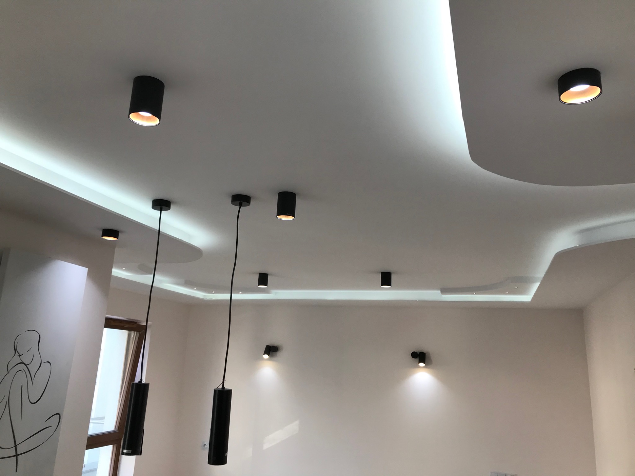 Nowoczesny sufit podwieszany z oświetleniem punktowym i wiszącymi lampami cylindrycznymi, minimalistyczny rysunek na ścianie w tle.