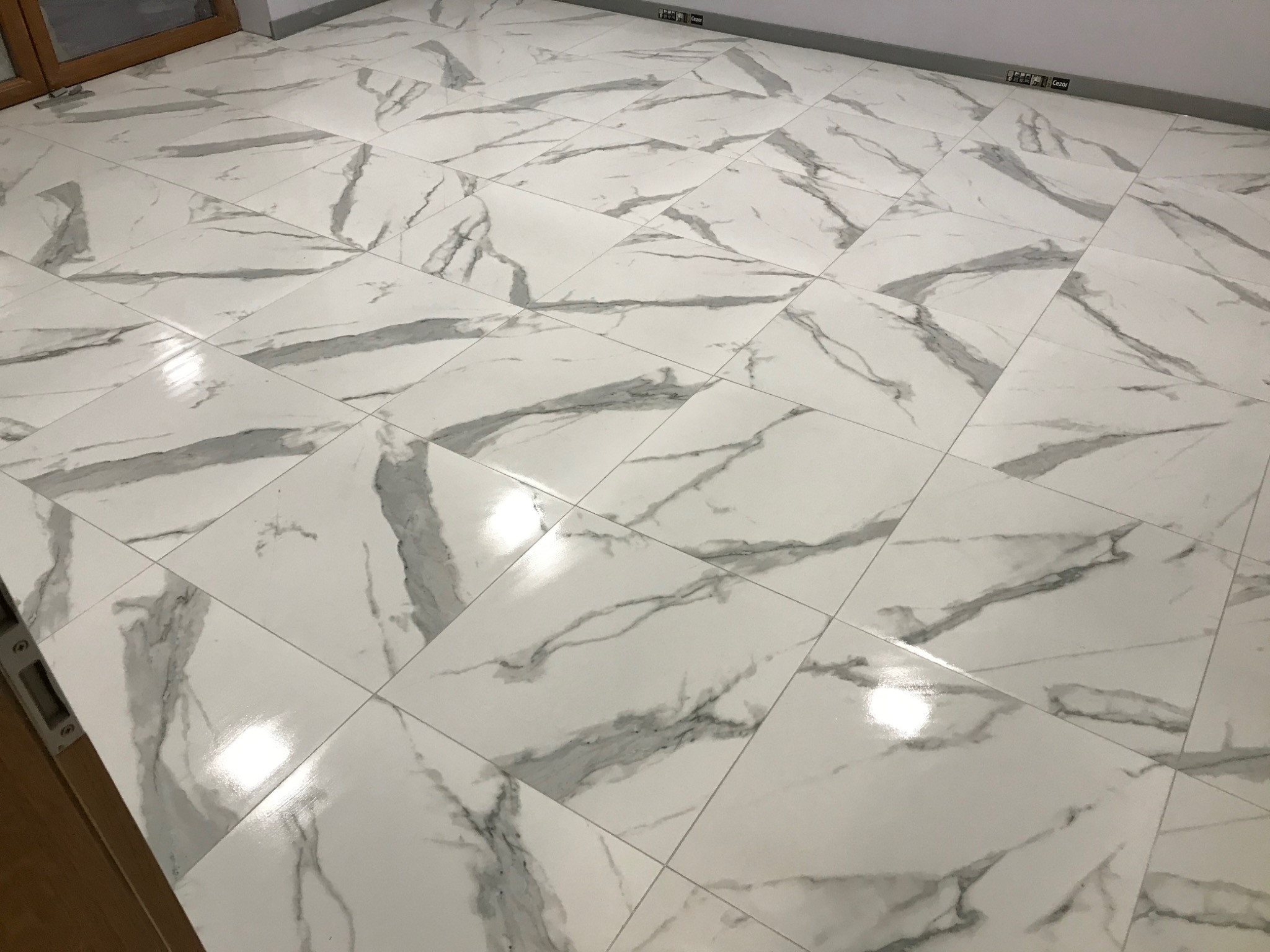Białe, błyszczące płytki podłogowe imitujące marmur z szarymi żyłkami, ułożone na podłodze w kwadraty, widoczne przy listwie przypodłogowej i fragmencie ościeżnicy drzwiowej.