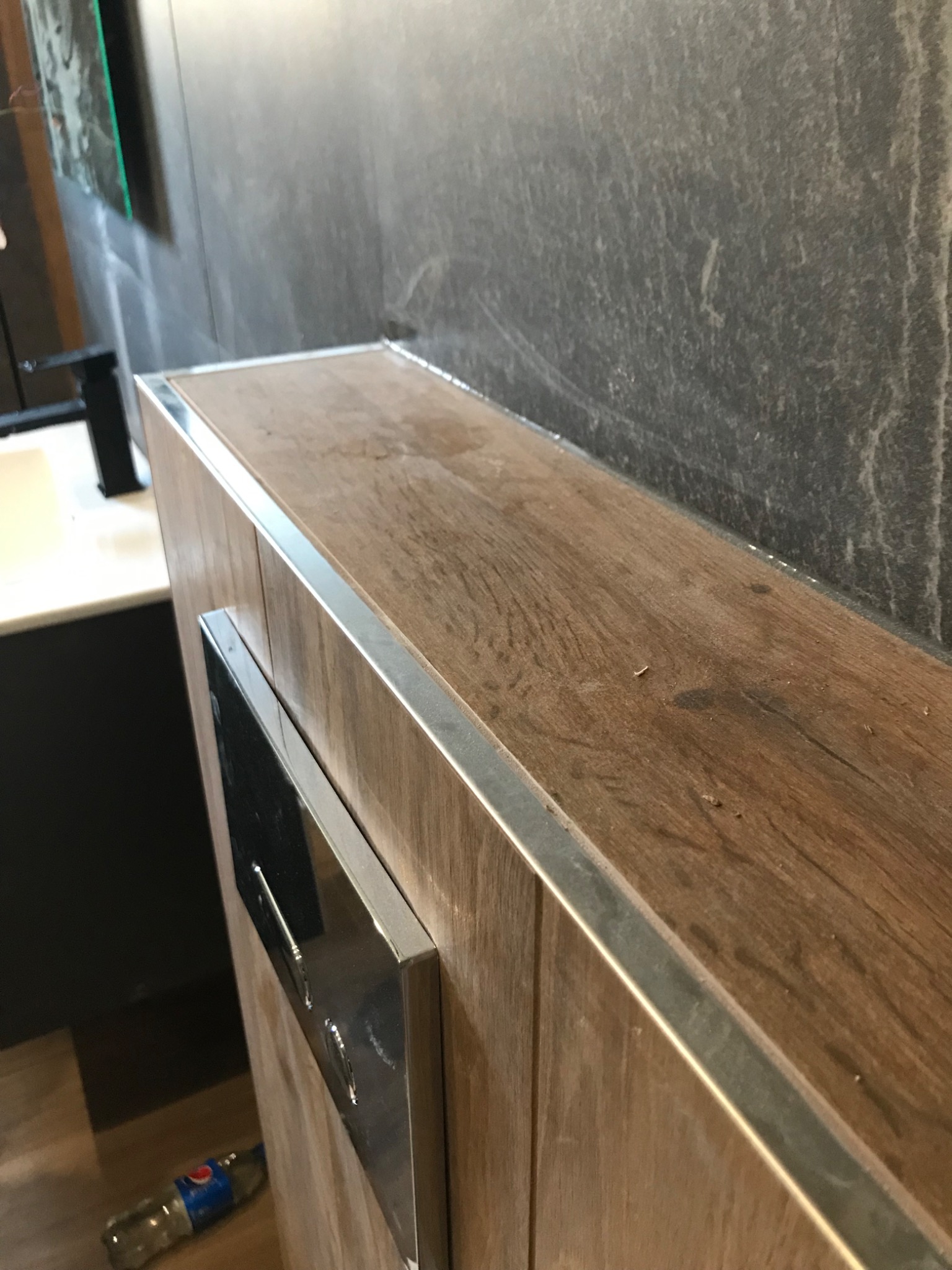 Drewnopodobna zabudowa toalety z chromowanym przyciskiem spłukiwania i obramowaniem, na tle szarej ściany z fakturą kamienia, z widoczną butelką wody na podłodze.