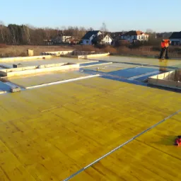 Budowa domu w trakcie realizacji: widok na żółtą, drewnianą płytę fundamentową z hydroizolacją, drewniane szalunki i pracownika z miotłą na tle zabudowy jednorodzinnej i lasu.