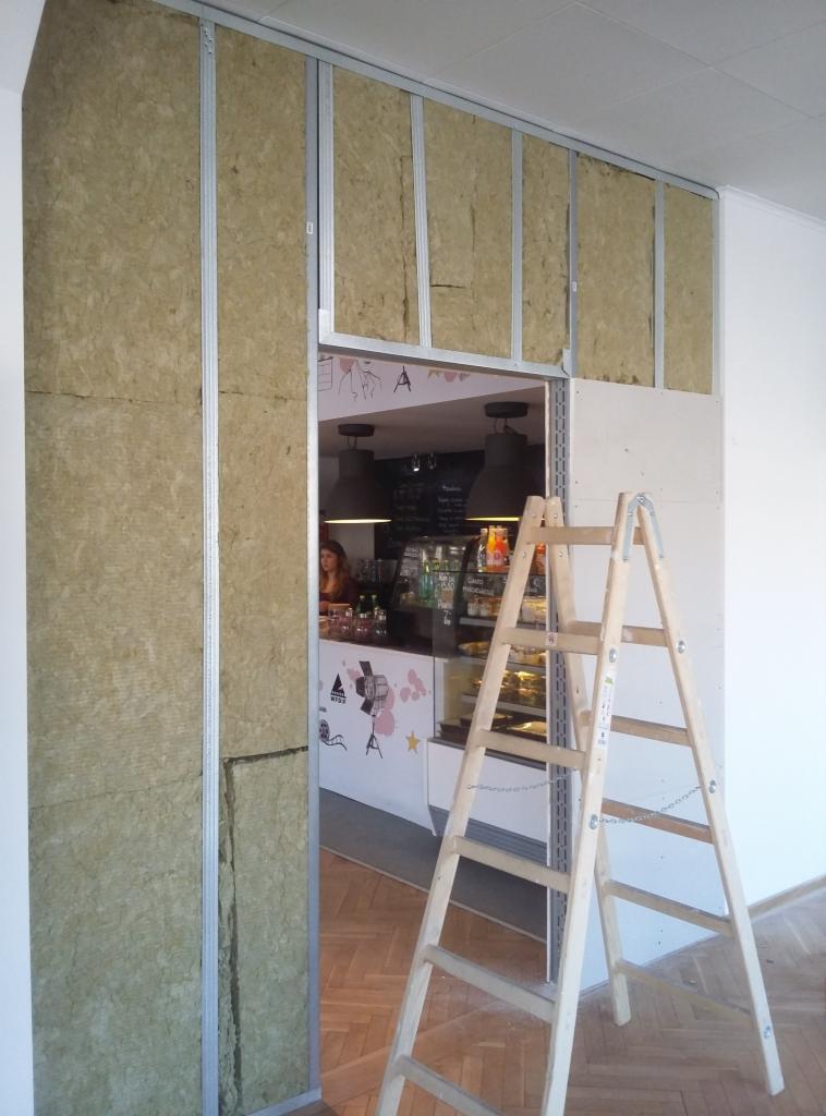 Montaż ścianki działowej z płyt kartonowo-gipsowych z izolacją z wełny mineralnej, widoczny metalowy szkielet i drabina, w tle wnętrze kawiarni.