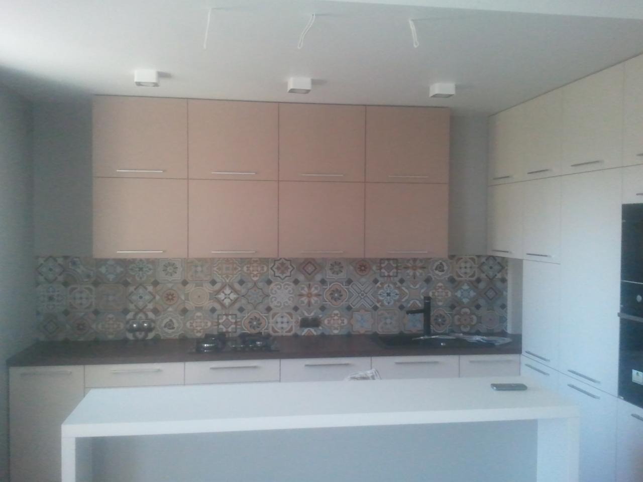 Nowoczesna kuchnia z beżowymi szafkami górnymi, białymi dolnymi i wyspą, oraz dekoracyjną mozaiką nad blatem.