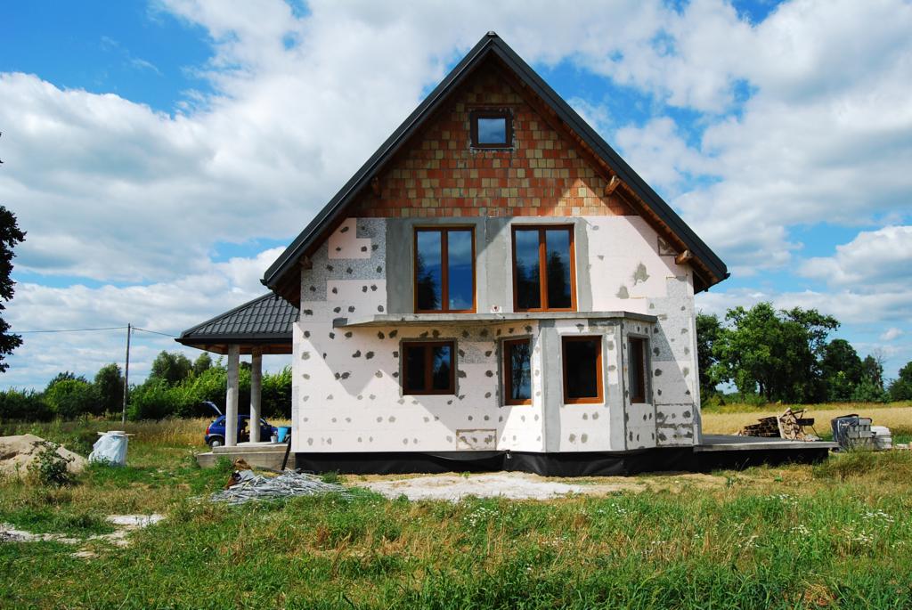 Dom jednorodzinny w Otwocku w trakcie ocieplania ścian zewnętrznych styropianem, widoczne okna z brązowymi ramami, fragment dachu z cegły oraz podbitka dachowa.