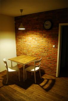Ceglana ściana w pokoju z drewnianą podłogą, oświetlona lampą wiszącą nad małym stołem z dwoma krzesłami; na ścianie widoczny zegar.