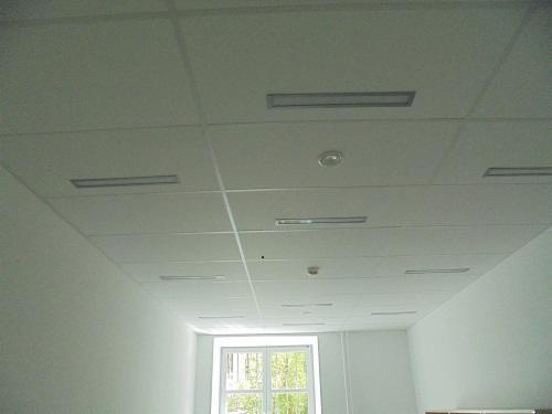 Kasetonowy sufit podwieszany w biurze z wbudowanymi liniowymi oprawami oświetleniowymi i czujnikiem dymu, widok z dołu na tle okna.