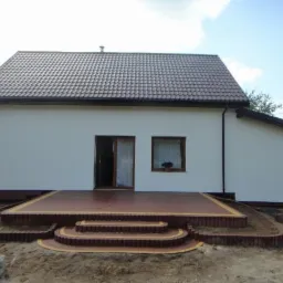 Nowy taras z kostki brukowej w odcieniach brązu i beżu, prowadzący do drzwi domu z białymi ścianami i brązowym dachem. Trzy stopnie z zaokrąglonymi krawędziami, ziemia wokół w trakcie prac...