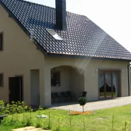 Gotowy dom z ciemną dachówką i beżową elewacją, widok z ogrodu z elementami grilla. Na dachu okno dachowe i komin.