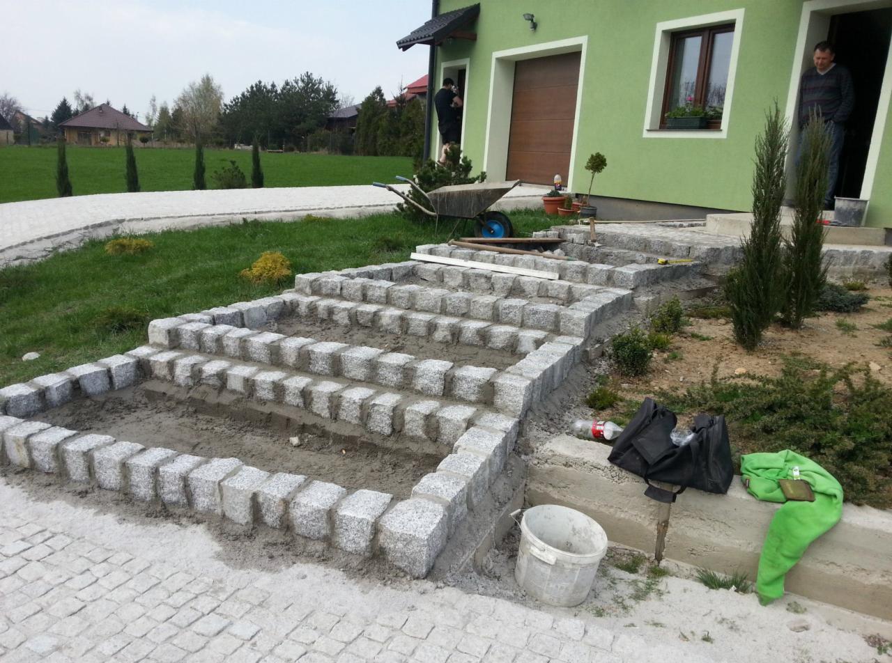 Budowa schodów zewnętrznych z kostki granitowej, widoczne etapy prac: ułożone obrzeża z kostki, wypełnienie ziemią, w tle dom z zieloną elewacją, podjazd z kostki, taczka i narzędzia.