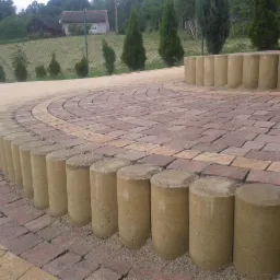 Okrągła ścieżka z kostki brukowej w odcieniach brązu i beżu, obramowana betonowymi słupkami. W tle trawnik, drzewa i dom.