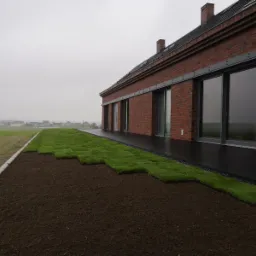 Układanie trawy z rolki przy nowoczesnym, ceglanym domu w pochmurny dzień. Widoczny fragment ogrodu z przygotowaną ziemią i ułożonymi fragmentami trawy w rolkach, przylegający do ciemnego tarasu.