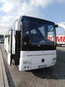 Biały autobus turystyczny marki Mercedes z otwartymi drzwiami, zaparkowany na placu z widoczną przyczepą w tle.