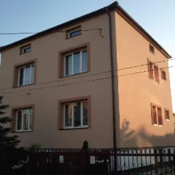 Dwupiętrowy dom z beżową elewacją i brązowymi obramowaniami okien, widoczny z zewnątrz, otoczony zielenią i ogrodzeniem.