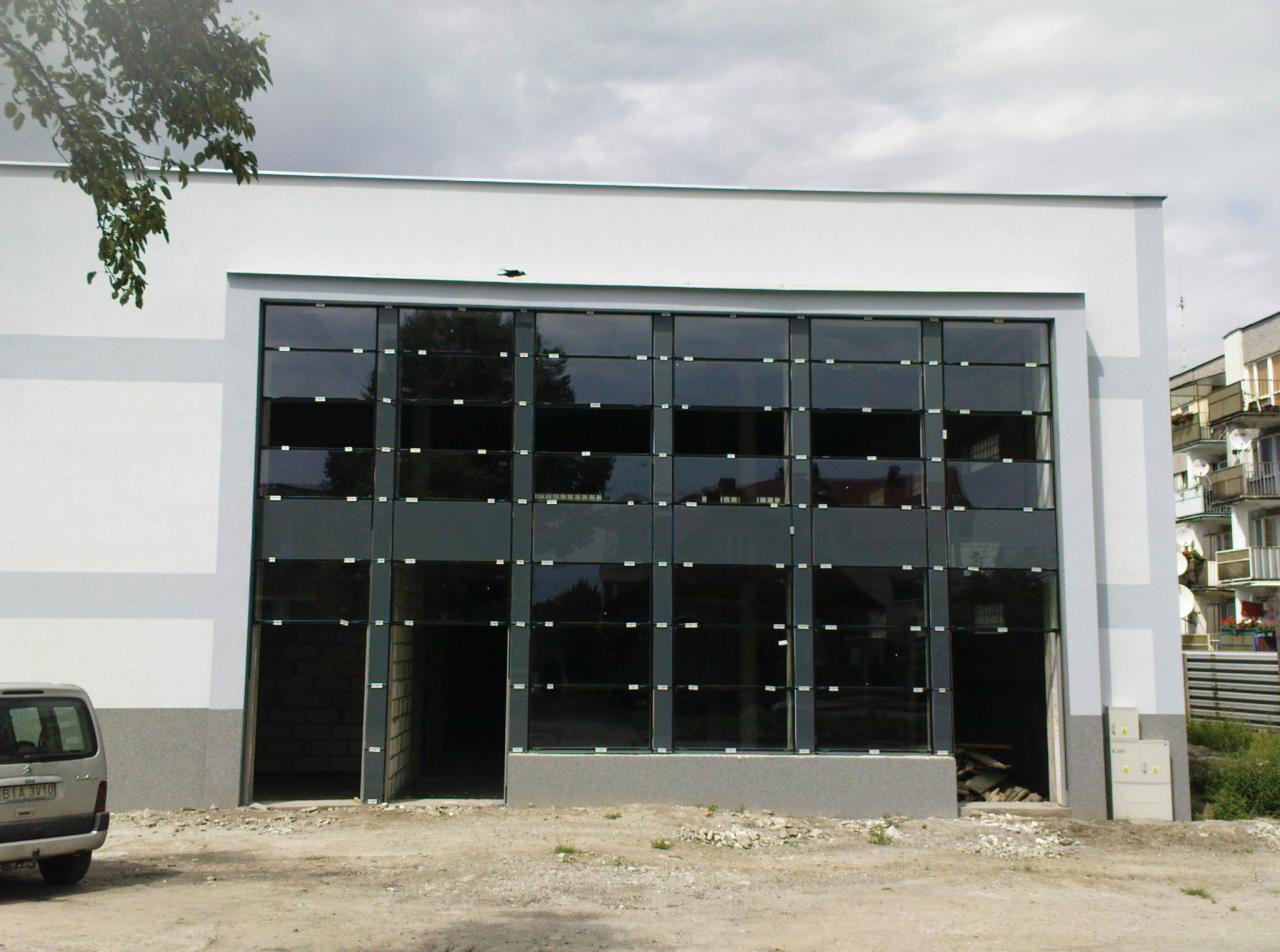 Nowoczesna elewacja budynku z dużymi, ciemnymi oknami w kwadratowych ramach. Widoczny fragment samochodu i budynek w tle. Szara kolorystyka elewacji.