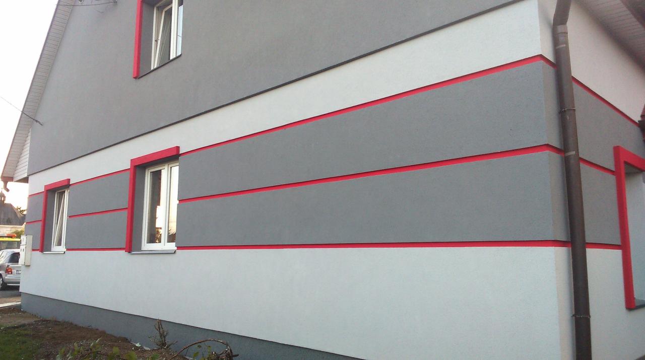 Dwukondygnacyjny dom po remoncie elewacji. Szara fasada z białymi pasami i czerwonymi akcentami wokół okien. Widok z zewnątrz, fragment budynku.