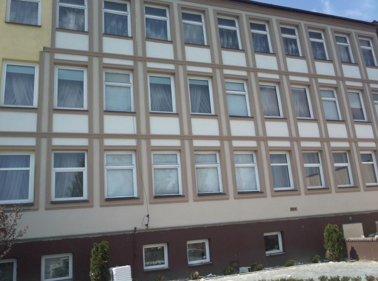 Wielopiętrowy budynek po renowacji elewacji, z widocznymi oknami z białymi ramami i kremowo-brązową elewacją. Dolna część budynku pomalowana na brązowo.