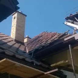 Demontaż dachówki przy kominie z podnośnika koszowego. Widoczne rynny, fragment dachu i komin z cegły. Prace dekarskie w toku, słoneczny dzień.