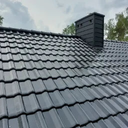 Błyszczące, grafitowe dachówki ceramiczne na skośnym dachu z kominem w tle pochmurnego nieba i drzew