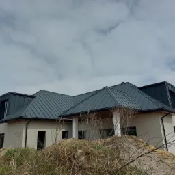 Nowoczesny dom z ciemnoszarą, metalową konstrukcją dachową, widoczny z perspektywy ziemi z usypanym kopcem ziemi na pierwszym planie, podczas prac wykończeniowych.