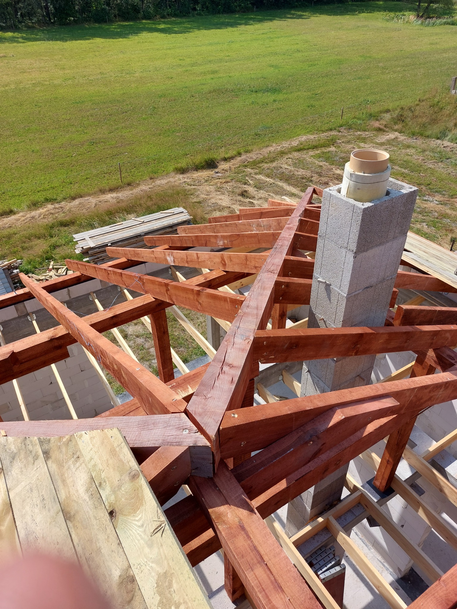 Widok z góry na drewnianą konstrukcję dachu w trakcie budowy, z kominem z szarych bloczków betonowych, na tle zielonego pola i lasu.