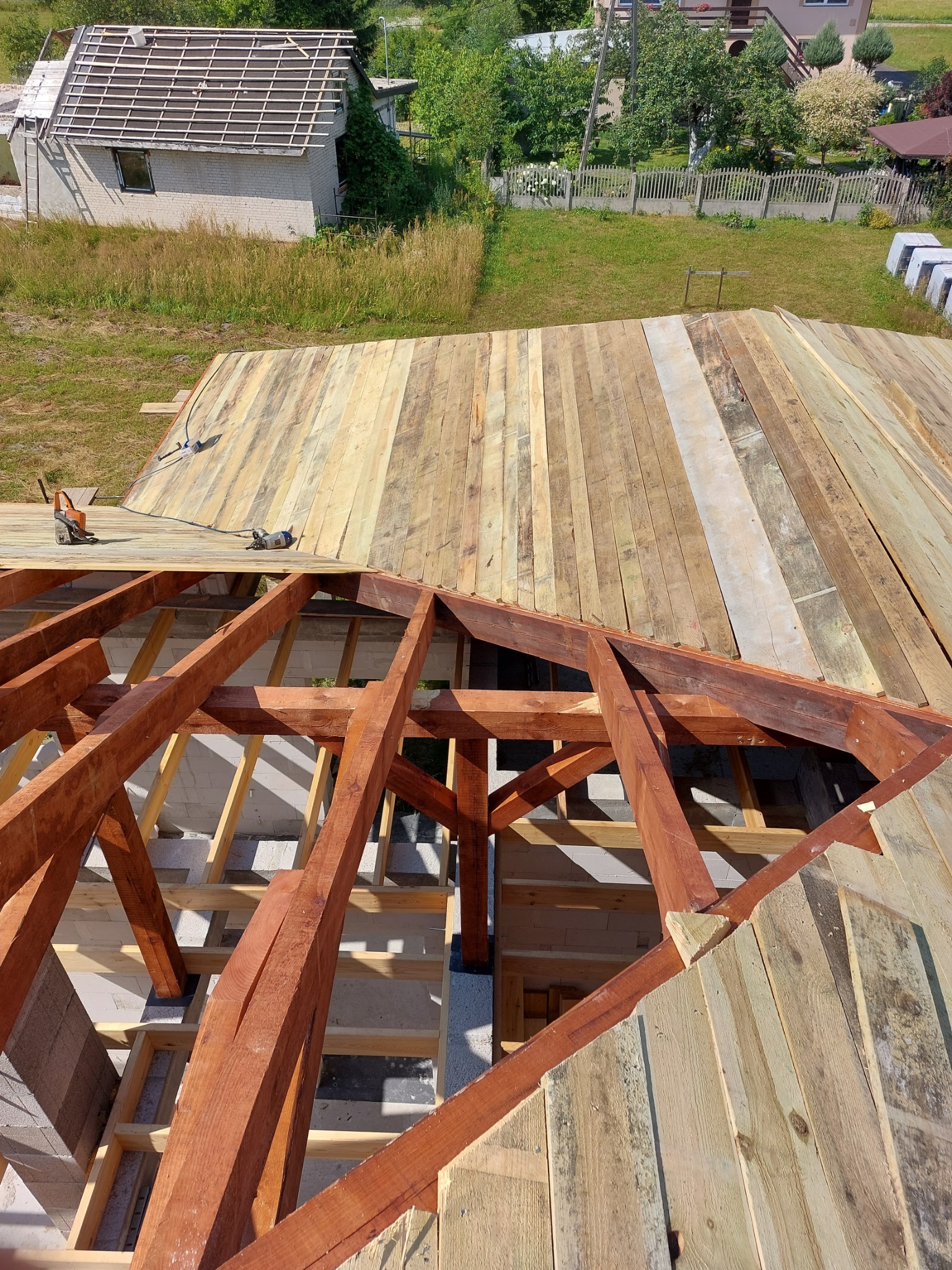 Konstrukcja dachu w trakcie budowy z widoczną więźbą i deskowaniem, narzędzia dekarskie na deskach, w tle budynek gospodarczy i zieleń.