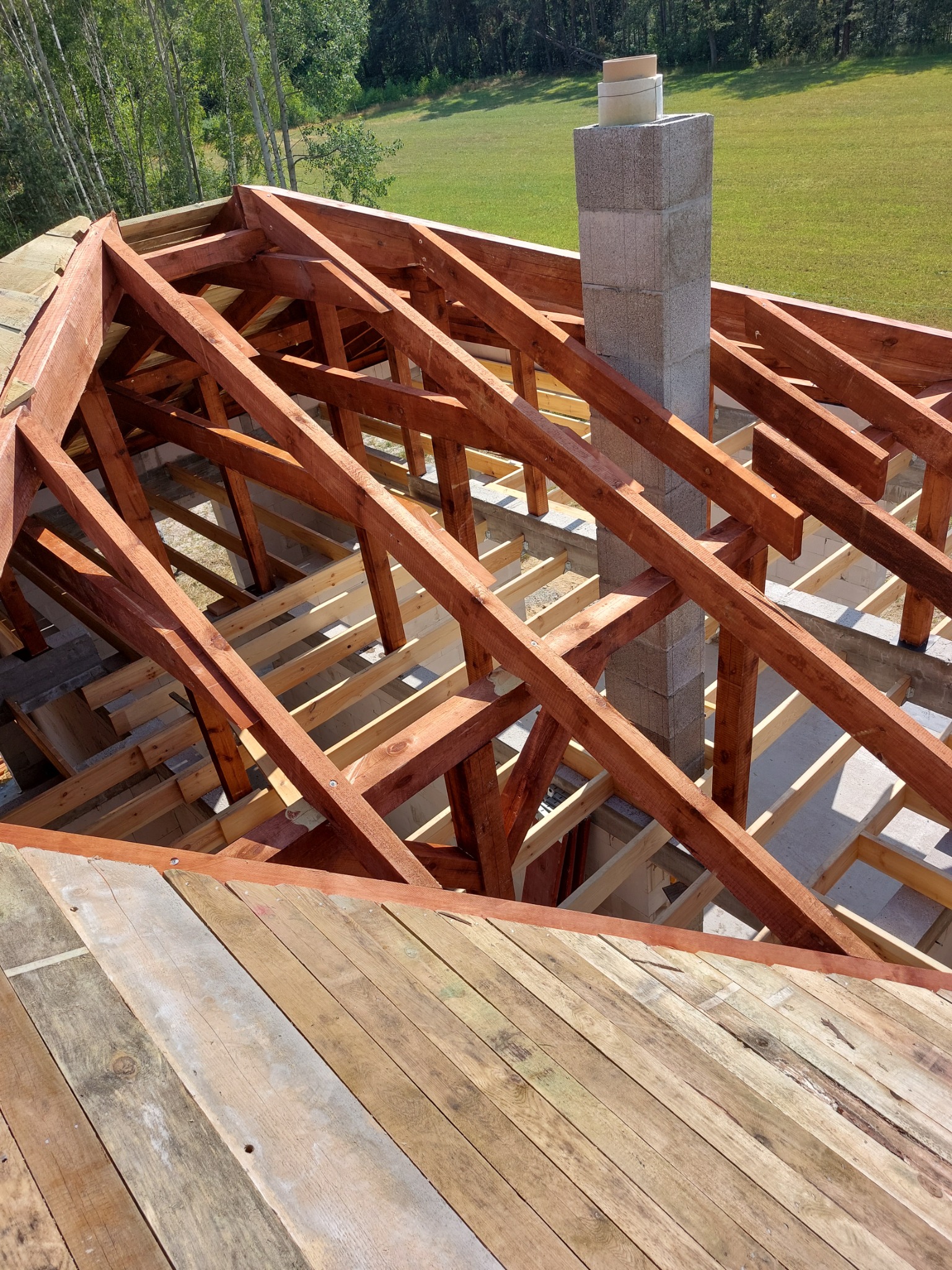 Konstrukcja drewniana dachu w trakcie budowy, z widocznym kominem z szarych bloczków i fragmentem poszycia z desek, na tle zielonego trawnika i drzew.