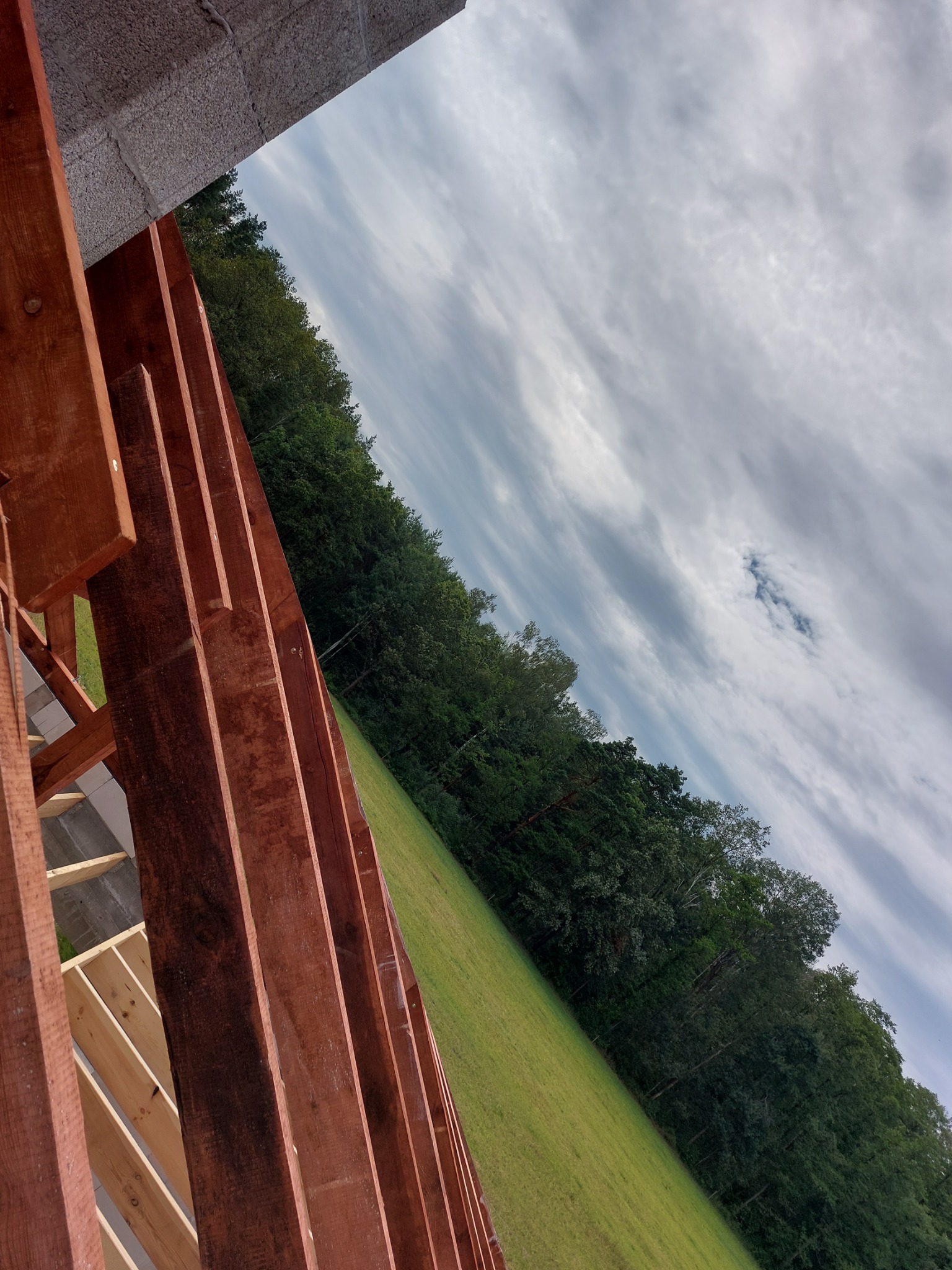 Fragment konstrukcji drewnianej dachu w trakcie budowy, widok na łąkę i pochmurne niebo z perspektywy dekarza.