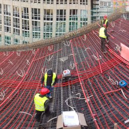 BKT TECHNIC MAREK WŁODARCZYK - Montaż rur w systemie rozprowadzenia ciepła pod betonową wylewkę, widoczni pracownicy w odblaskowych kamizelkach i kaskach, w tle budynek w trakcie budowy.