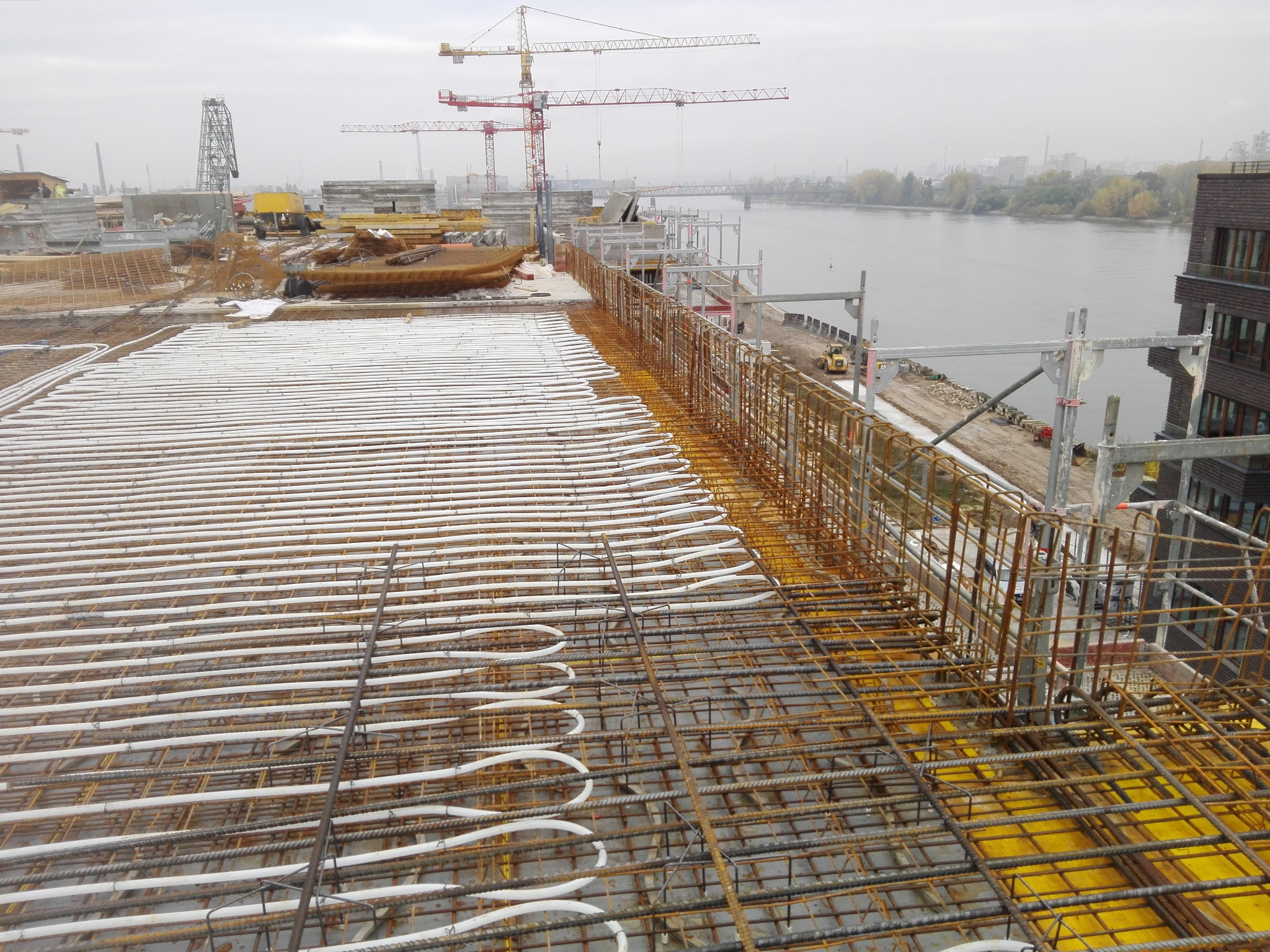 Białe rury systemu grzewczego ułożone w regularnych odstępach na metalowej siatce zbrojeniowej na budowie, w tle dźwigi i rzeka.