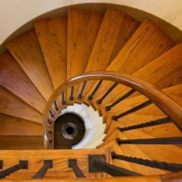 Drewniane schody spiralne widziane z góry, z ciemnymi metalowymi balustradami i okrągłym oknem w tle.