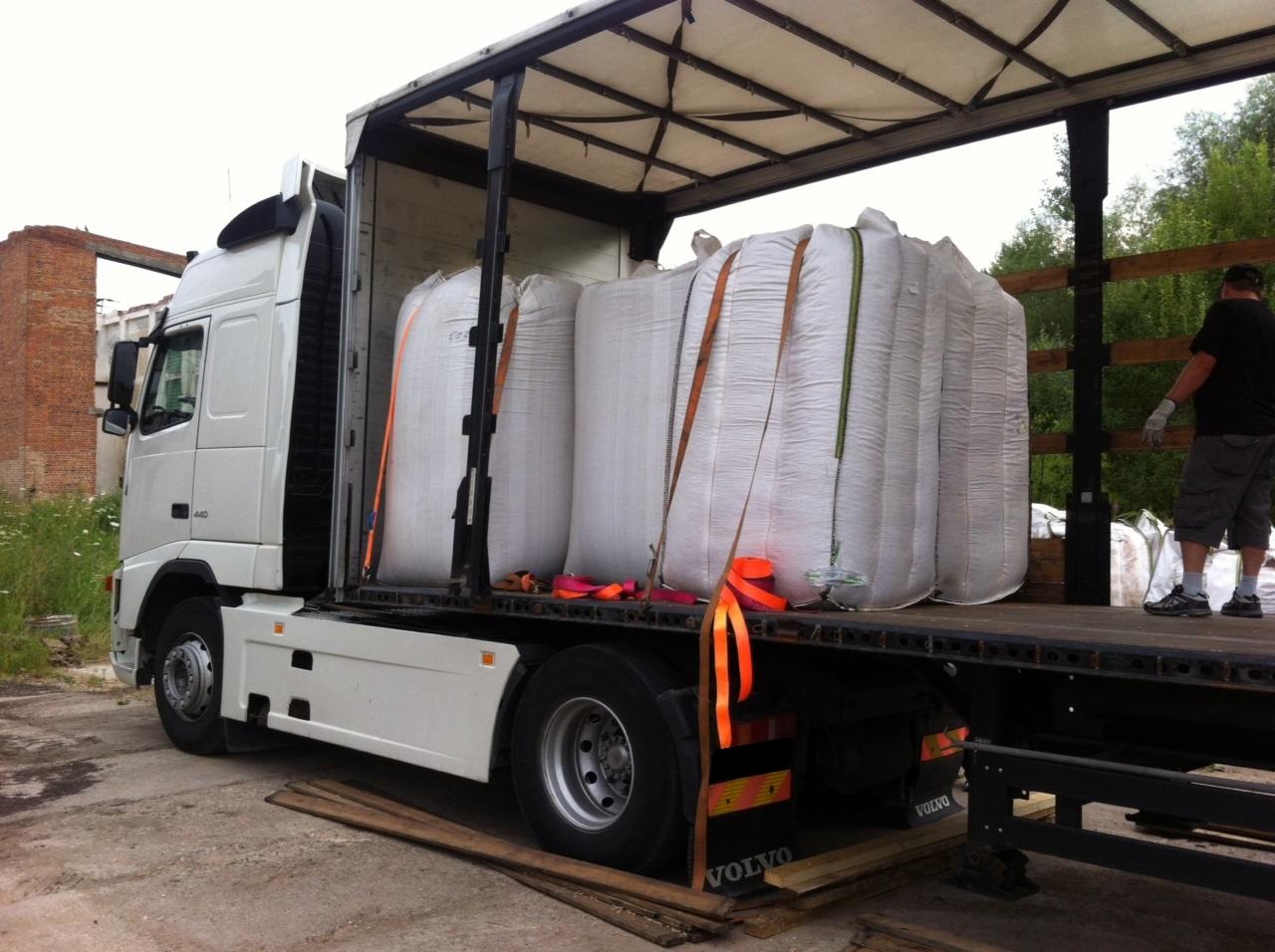 Ciężarówka Volvo z naczepą, na której załadowane są duże, białe worki z pelletem, zabezpieczone pomarańczowymi pasami transportowymi. W tle widoczny fragment ceglanego budynku i zieleń.