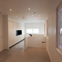 Wnętrze w trakcie wykańczania: białe ściany, gładka szara podłoga, zabudowa meblowa z miejscem na telewizor, widoczne okablowanie na suficie, minimalistyczny styl.