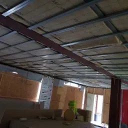 Montaż izolacji z wełny mineralnej w metalowym stelażu pod stropem, widoczne płyty OSB na ścianach i materiały budowlane w pomieszczeniu.