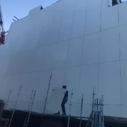 Montaż białych paneli elewacyjnych na rusztowaniu, widoczny dźwig budowlany w tle.