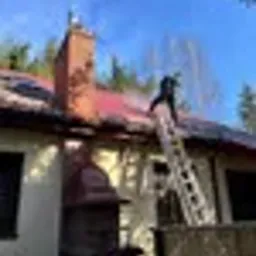Pracownik na drabinie czyści dach domu z czerwonej dachówki, widoczny komin i zieleń drzew w tle, jasne niebo.