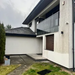 Dwupiętrowy dom z szarą elewacją i szklanym balkonem. Widoczne ślady zabrudzeń na elewacji w dolnej części budynku. Kostka brukowa przed wejściem.