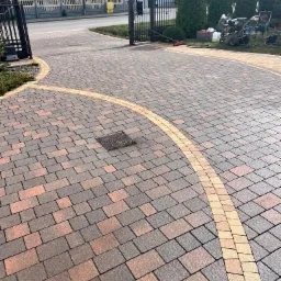 Czysta kostka brukowa w odcieniach brązu i beżu, ułożona w geometryczny wzór. W tle brama, zieleń i sprzęt do czyszczenia pod ciśnieniem.