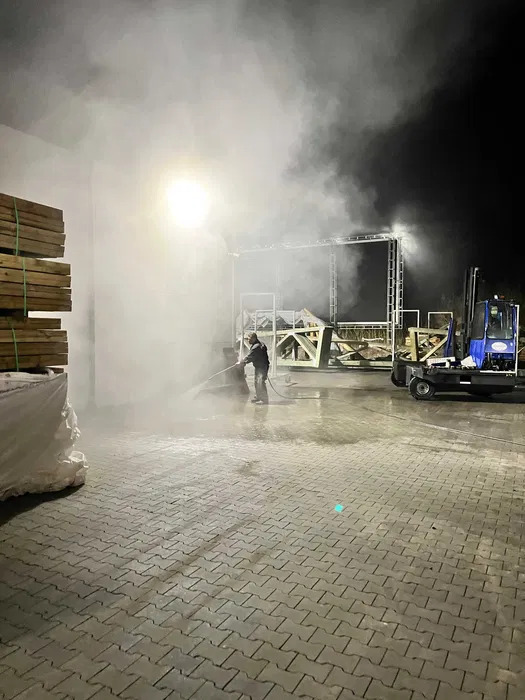 Czyszczenie kostki brukowej pod silnym ciśnieniem, z widocznym efektem pary wodnej i stojącym w tle wózkiem widłowym. Praca wykonywana nocą.