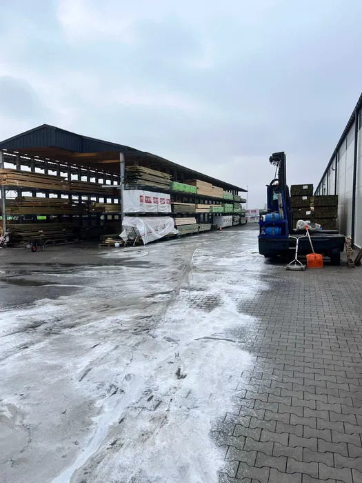 Magazyn z materiałami budowlanymi i wózkiem widłowym na zewnątrz. Część kostki brukowej jest umyta, widoczne ślady po czyszczeniu. Pochmurny dzień.