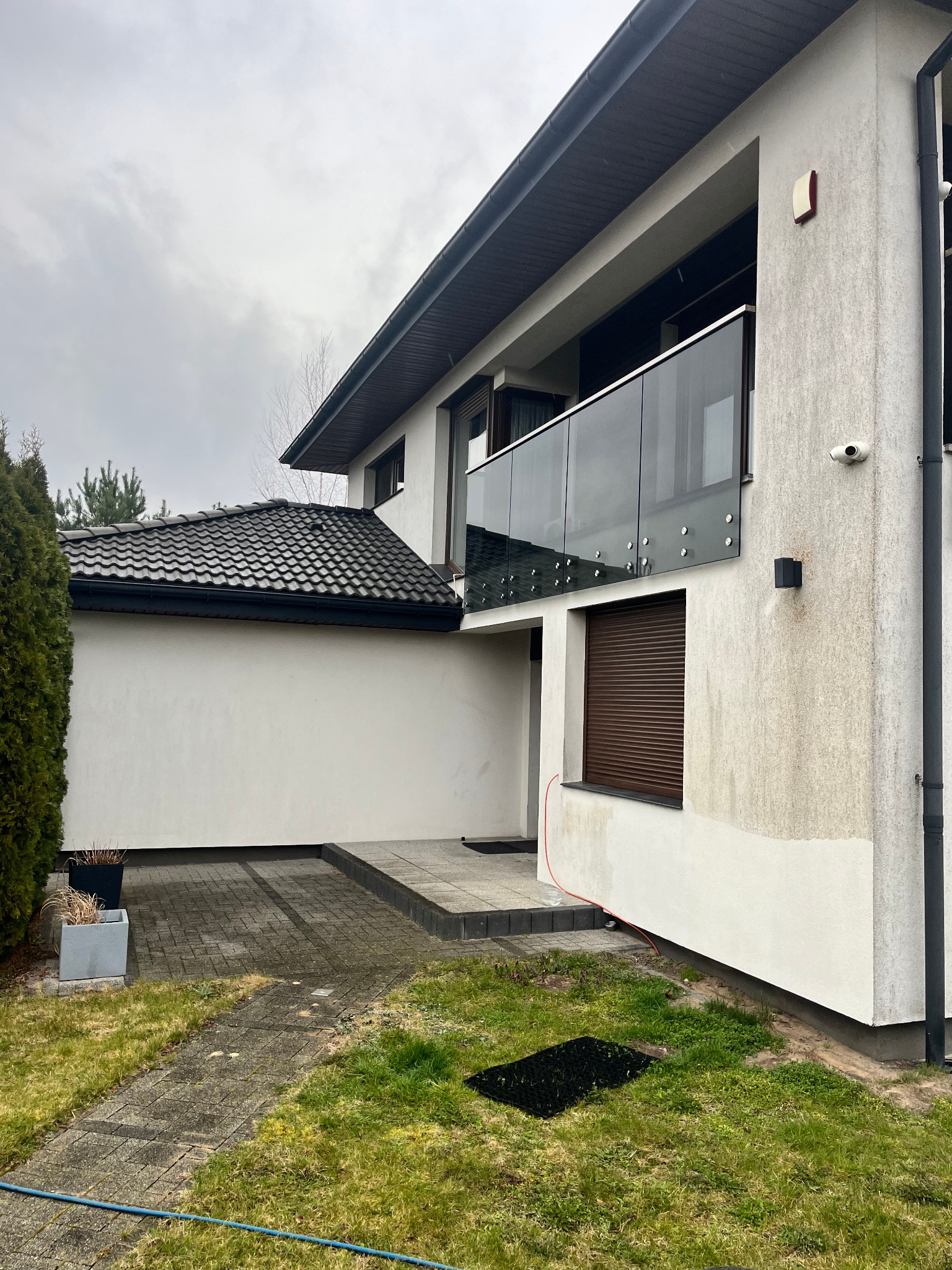 Dwupiętrowy dom z szarą elewacją i szklanym balkonem. Widoczne ślady zabrudzeń na elewacji w dolnej części budynku. Kostka brukowa przed wejściem.