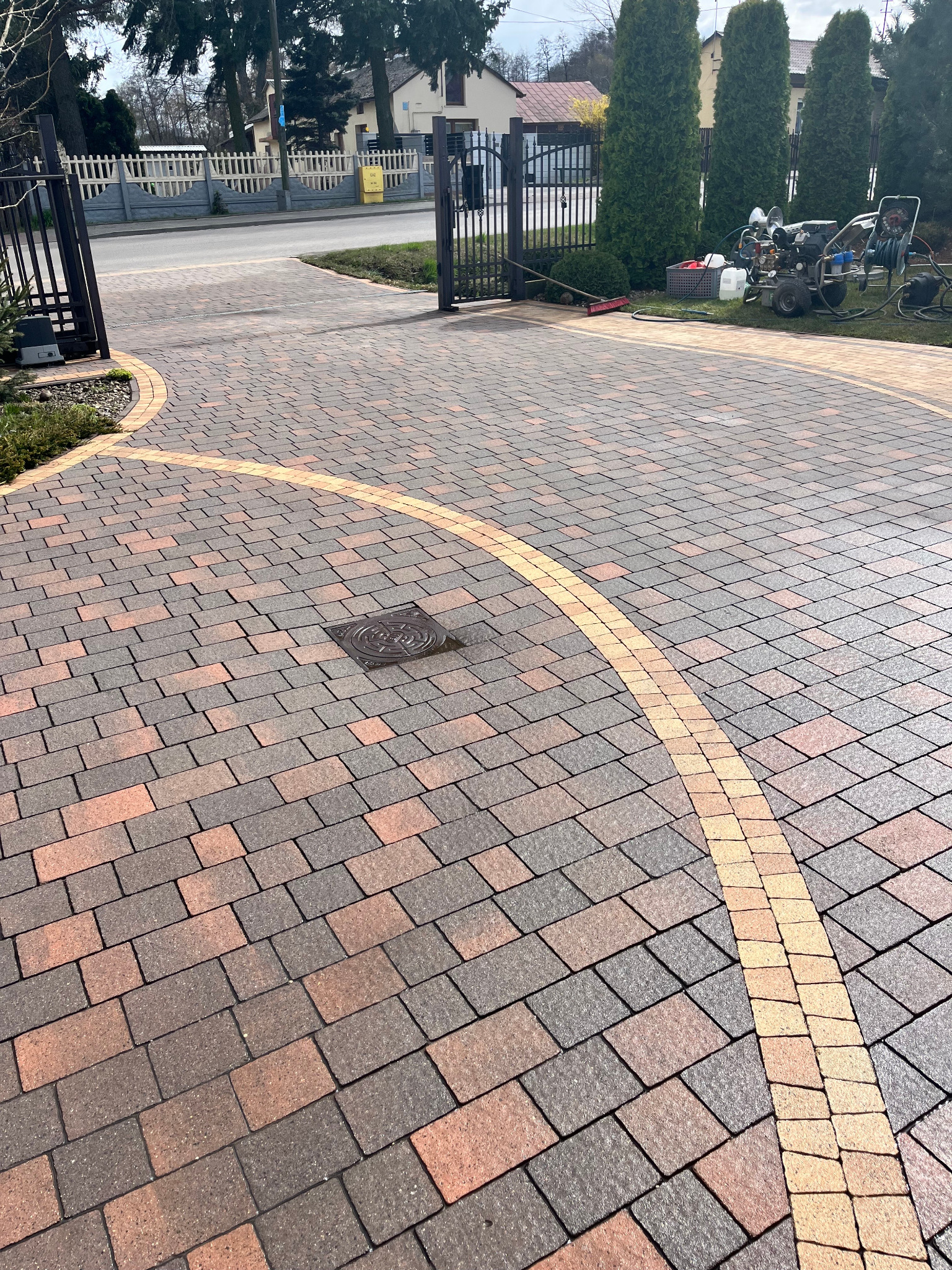 Czysta kostka brukowa w odcieniach brązu i beżu, ułożona w geometryczny wzór. W tle brama, zieleń i sprzęt do czyszczenia pod ciśnieniem.