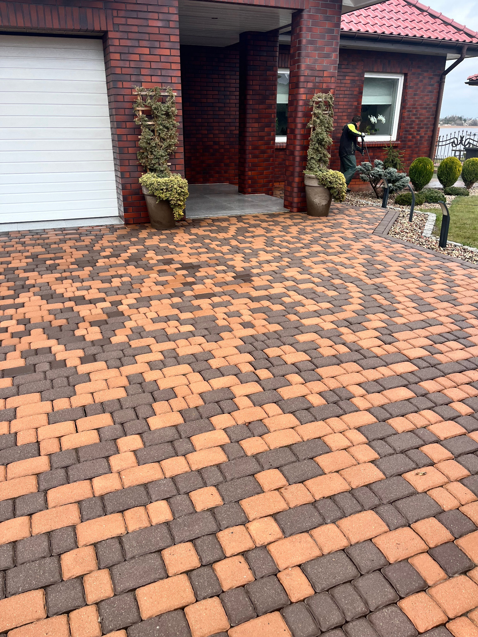 Dwukolorowa kostka brukowa przed domem z cegły, osoba w oddali czyści powierzchnię. Widoczne rośliny w donicach i elementy architektury ogrodowej.