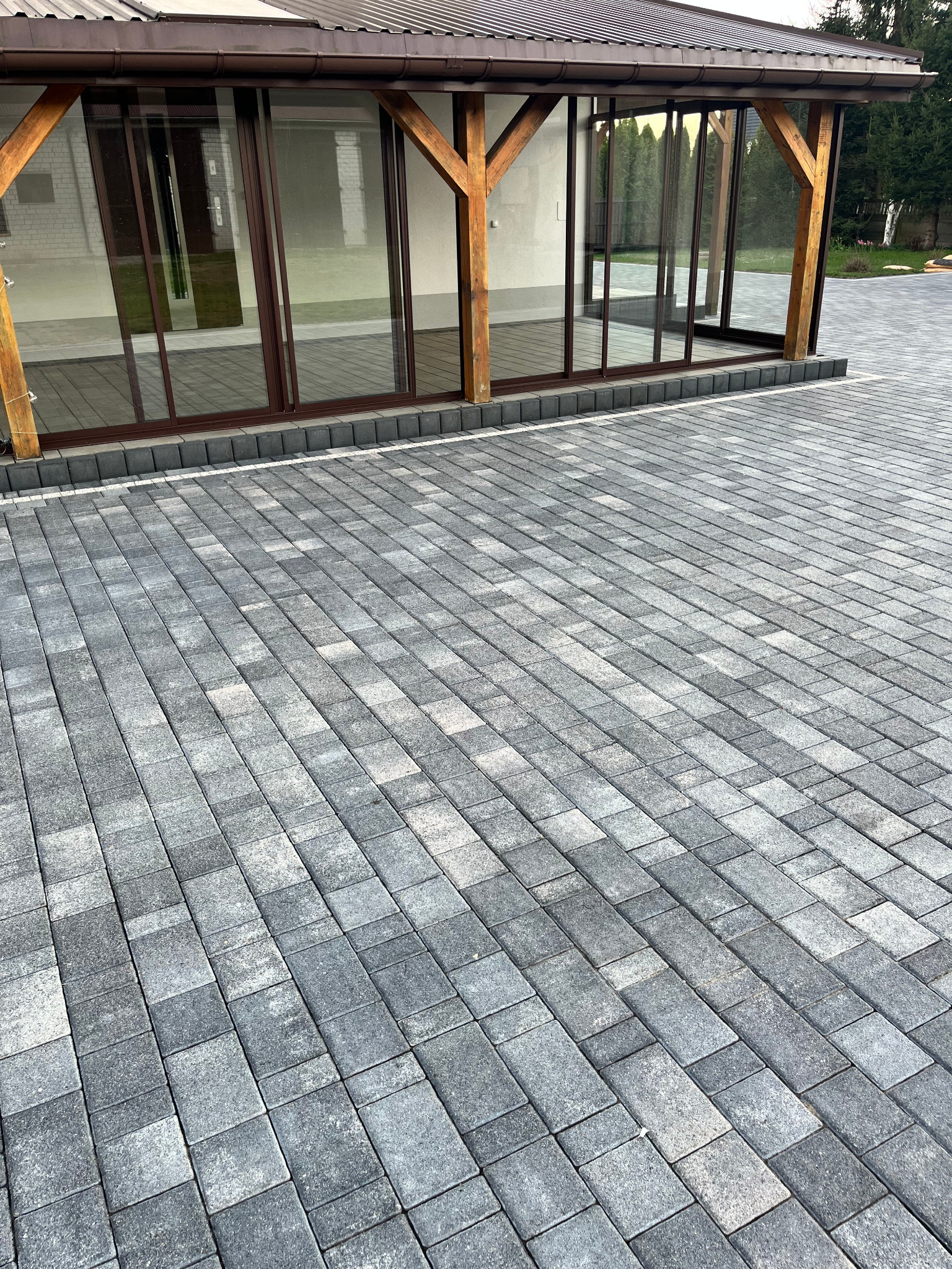 Szara kostka brukowa ułożona przed przeszkloną altaną z drewnianą konstrukcją. Widok z perspektywy ziemi, podkreślający wzór kostki.