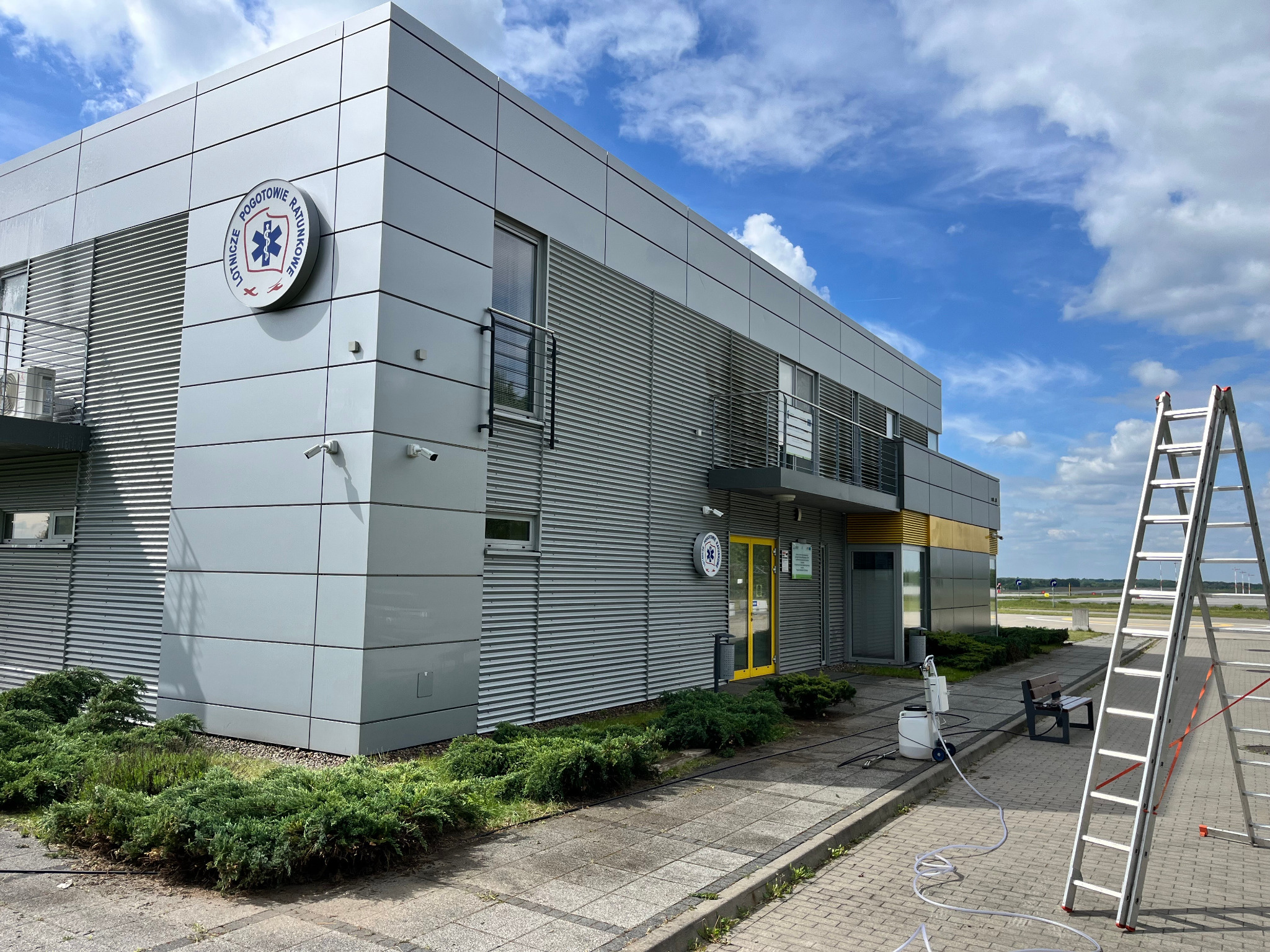 Szara elewacja budynku Lotniczego Pogotowia Ratunkowego z drabiną i sprzętem do czyszczenia na zewnątrz. Błękitne niebo w tle.