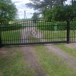 Czarna metalowa brama wjazdowa z ozdobnymi elementami w kolorze złotym, prowadząca na szutrową drogę wśród zieleni.