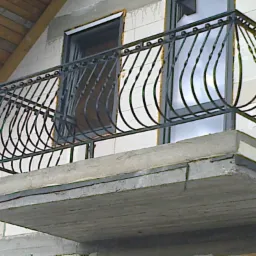 Czarna, metalowa balustrada balkonowa z dekoracyjnymi, zakrzywionymi prętami, zamontowana na surowym, betonowym balkonie w trakcie budowy domu z białego bloczka.