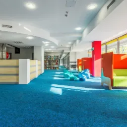 Jasne wnętrze biblioteki lub biura z intensywnie niebieską wykładziną dywanową, strefą relaksu z pufami i kabinami do pracy w skupieniu z pomarańczowymi ściankami i jasnozielonymi siedziskami.