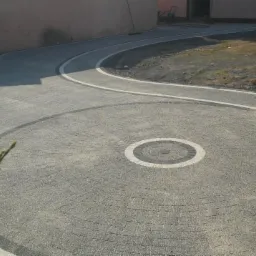 Szara kostka brukowa ułożona w okrągły wzór z białymi i ciemnoszarymi obwódkami, tworząca ścieżkę wijącą się przez ogród.