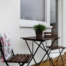 Zestaw bistro z ciemnego drewna i czarnego metalu na drewnianym tarasie przy białej ścianie budynku z dużym oknem; na krześle leży pled w kwiaty.